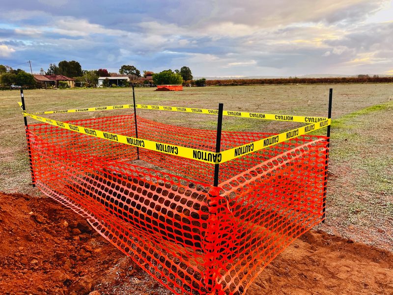Temporary orange safety fencing and caution tape set up around an open excavation on a rural block